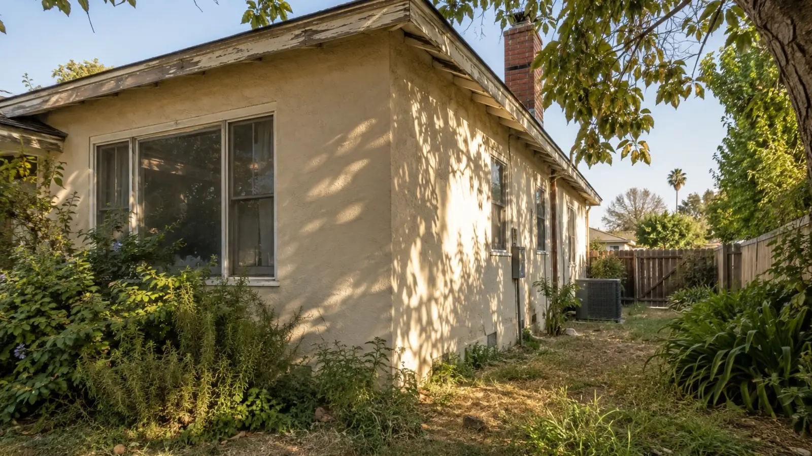 The exterior corner of an older stucco home showing an honestly weathered roofline and a slightly overgrown side yard, warm late afternoon light, mature tree casting shade, Riverside CA