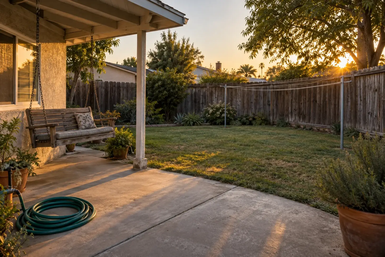 A small backyard at golden hour with an empty clothesline, a still porch swing, and a coiled hose by a weathered fence, Southern California