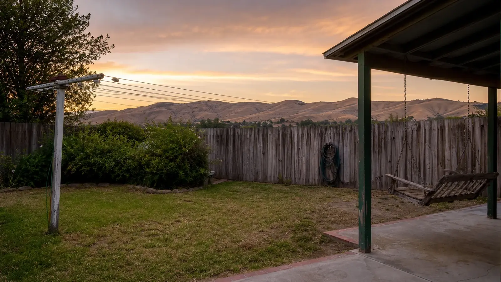 A still backyard at dusk with an empty clothesline, a quiet porch swing and a coiled garden hose against a weathered fence, gold hills beyond, Temecula CA