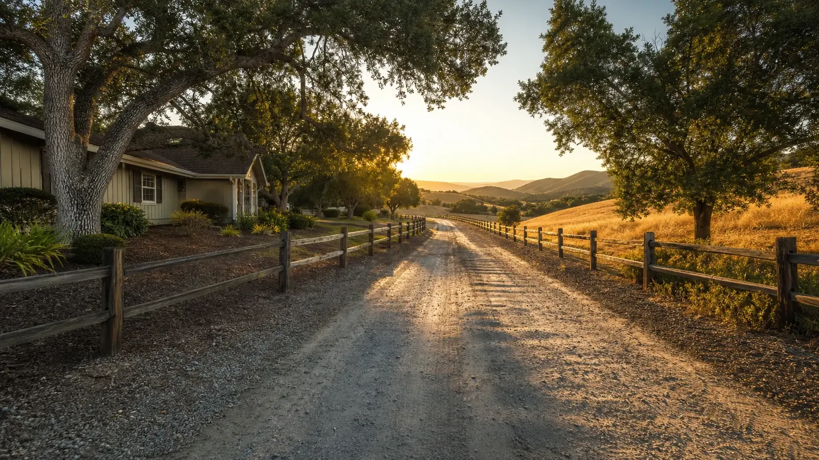 A long gravel driveway lined with split-rail fencing and mature oak trees leading toward gold foothills at sunset, Temecula CA