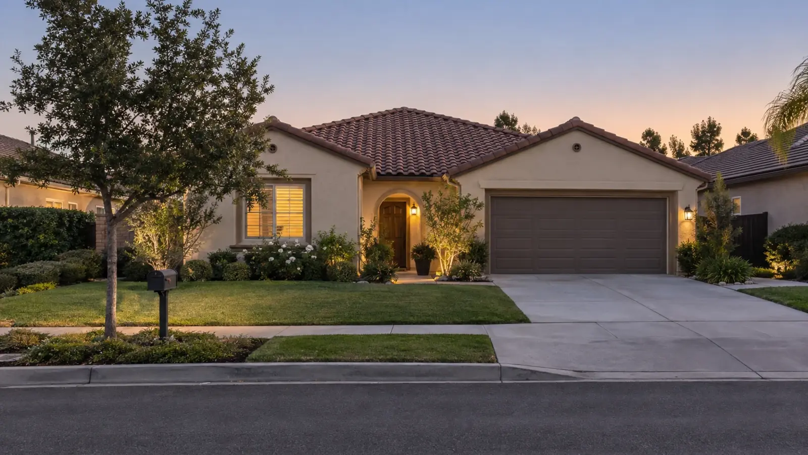 A modest stucco home with a Spanish tile roof seen from across a quiet street at twilight, one window lit and the porch light on, Murrieta CA