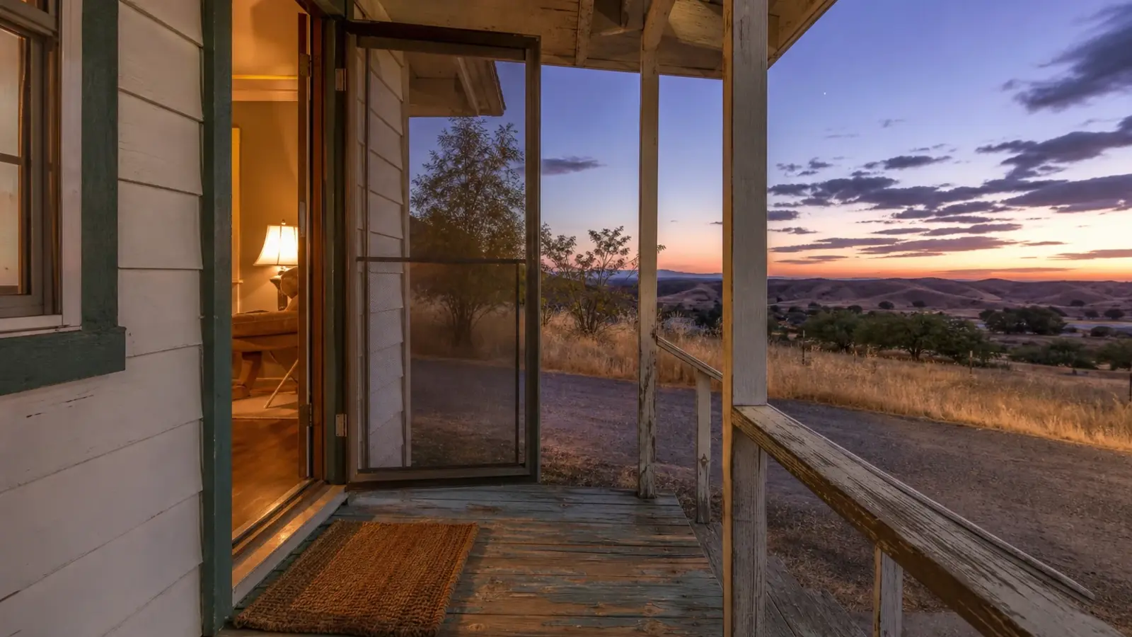 The open front door of a modest older ranch home glowing with lamplight at dusk, rural Southwest Riverside County CA